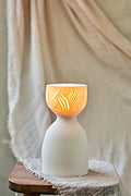 A ceramic translucent porcelain glowing lamp with a glowing warm light inside, featuring a carved pattern on the upper half and a smooth matte white lower body, displayed on a rustic wooden stool with draped fabric in the background.

