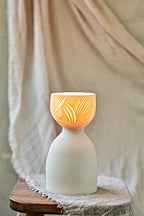 A ceramic translucent porcelain glowing lamp with a glowing warm light inside, featuring a carved pattern on the upper half and a smooth matte white lower body, displayed on a rustic wooden stool with draped fabric in the background.

