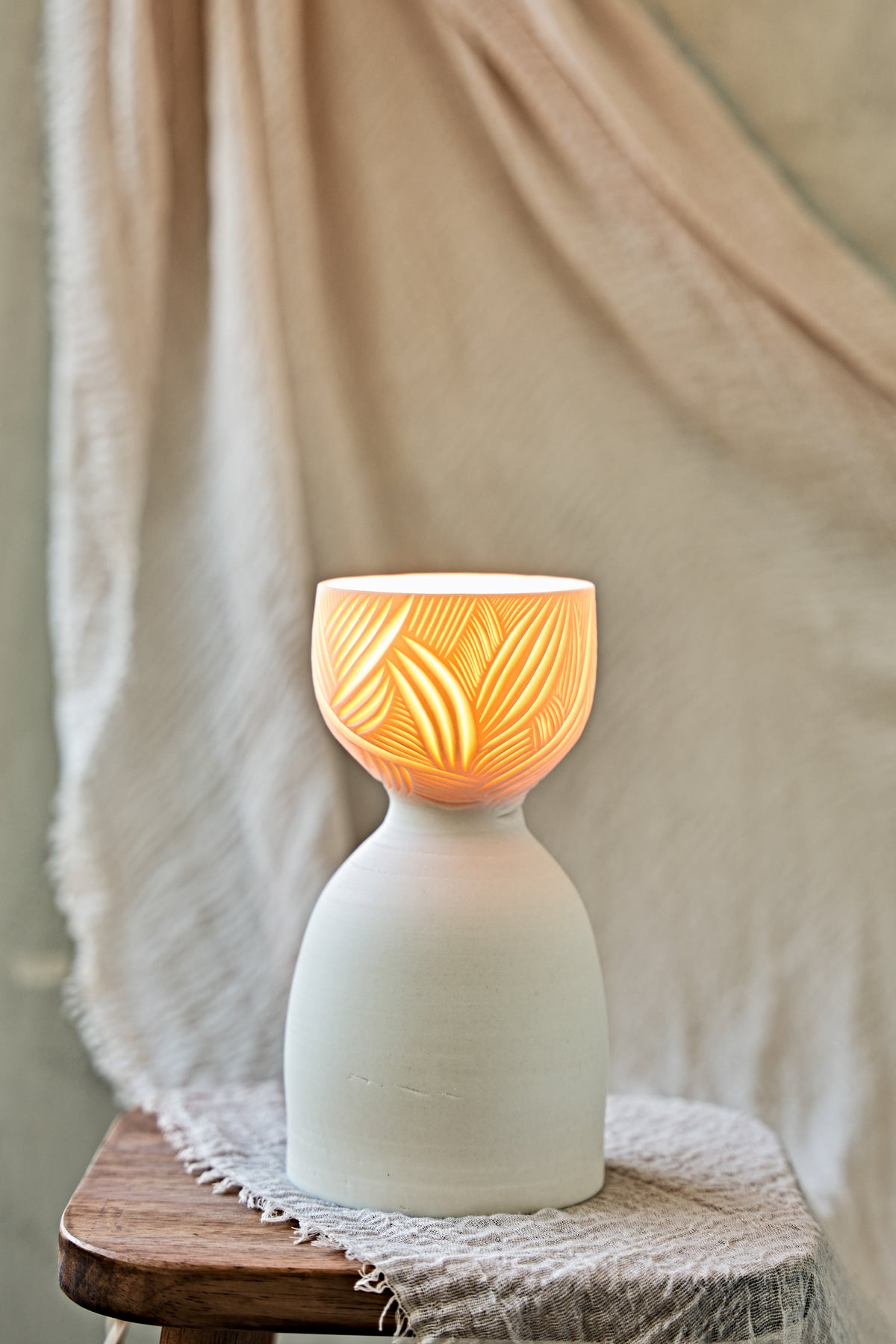 A ceramic translucent porcelain glowing lamp with a glowing warm light inside, featuring a carved pattern on the upper half and a smooth matte white lower body, displayed on a rustic wooden stool with draped fabric in the background.

