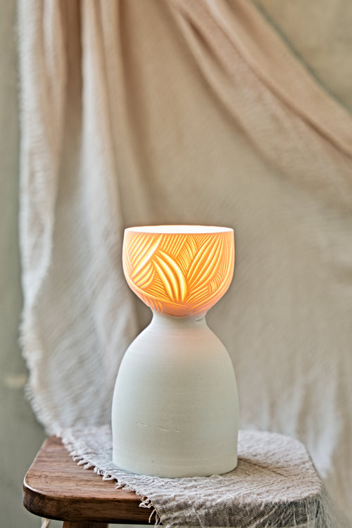 A ceramic translucent porcelain glowing lamp with a glowing warm light inside, featuring a carved pattern on the upper half and a smooth matte white lower body, displayed on a rustic wooden stool with draped fabric in the background.

