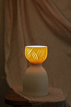 A ceramic translucent porcelain glowing lamp with a glowing amber warm light inside, featuring a carved pattern on the upper half and a smooth matte white lower body, displayed on a rustic wooden stool with draped fabric in the background.