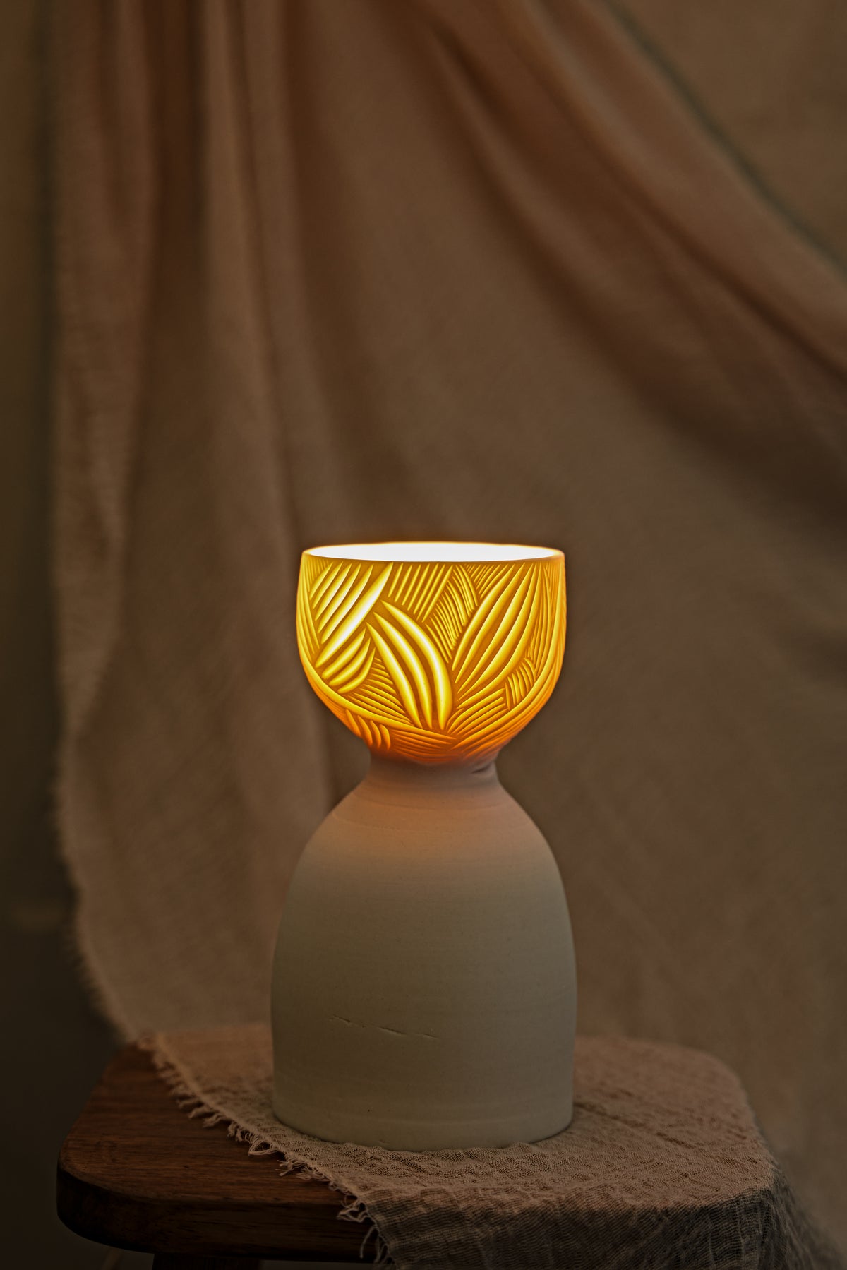 A ceramic translucent porcelain glowing lamp with a glowing amber warm light inside, featuring a carved pattern on the upper half and a smooth matte white lower body, displayed on a rustic wooden stool with draped fabric in the background.
