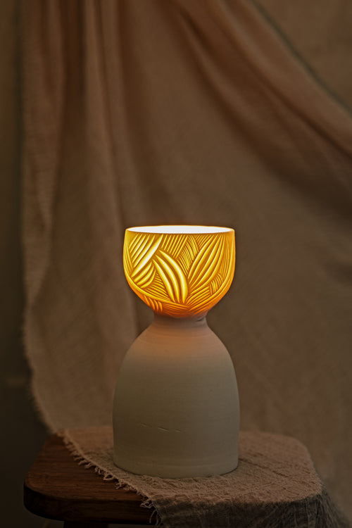 A ceramic translucent porcelain glowing lamp with a glowing amber warm light inside, featuring a carved pattern on the upper half and a smooth matte white lower body, displayed on a rustic wooden stool with draped fabric in the background.