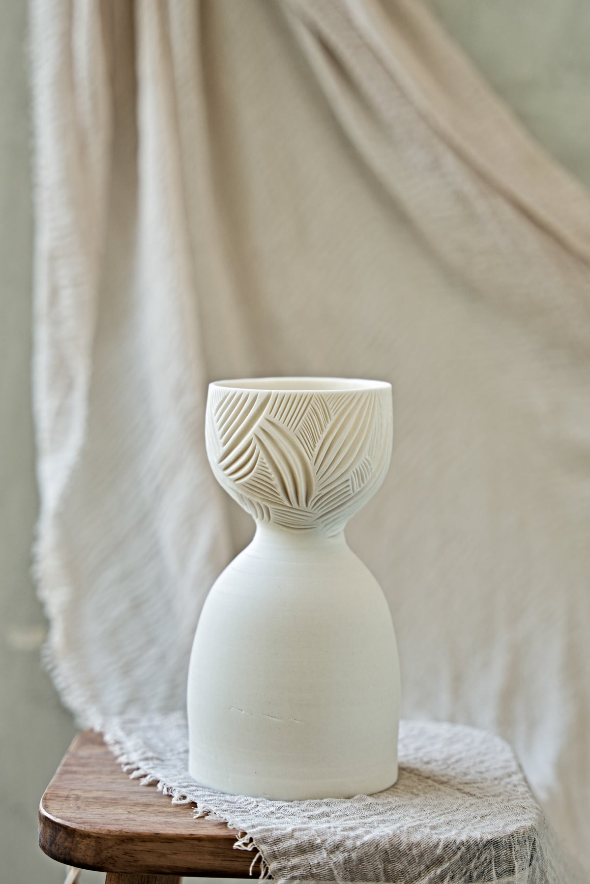 A ceramic translucent porcelain glowing lamp featuring a carved pattern on the upper half and a smooth matte white lower body, displayed on a rustic wooden stool with draped fabric in the background.