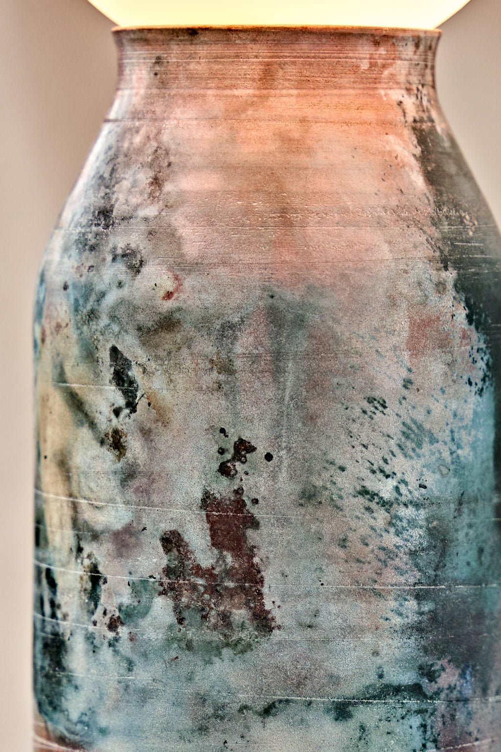 Close-up of a ceramic vase with a textured, distressed finish showing mottled shades of teal, brown, and copper-orange, with a light cream-colored top visible at the top edge.