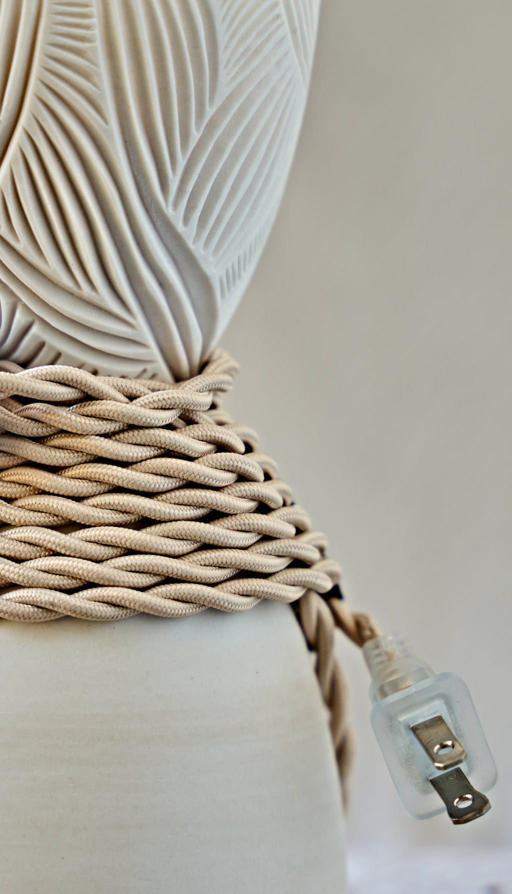 Close-up of a decorative lamp base wrapped in thick braided beige fabric cord, with a clear plastic plug visible against a soft light gray background.
