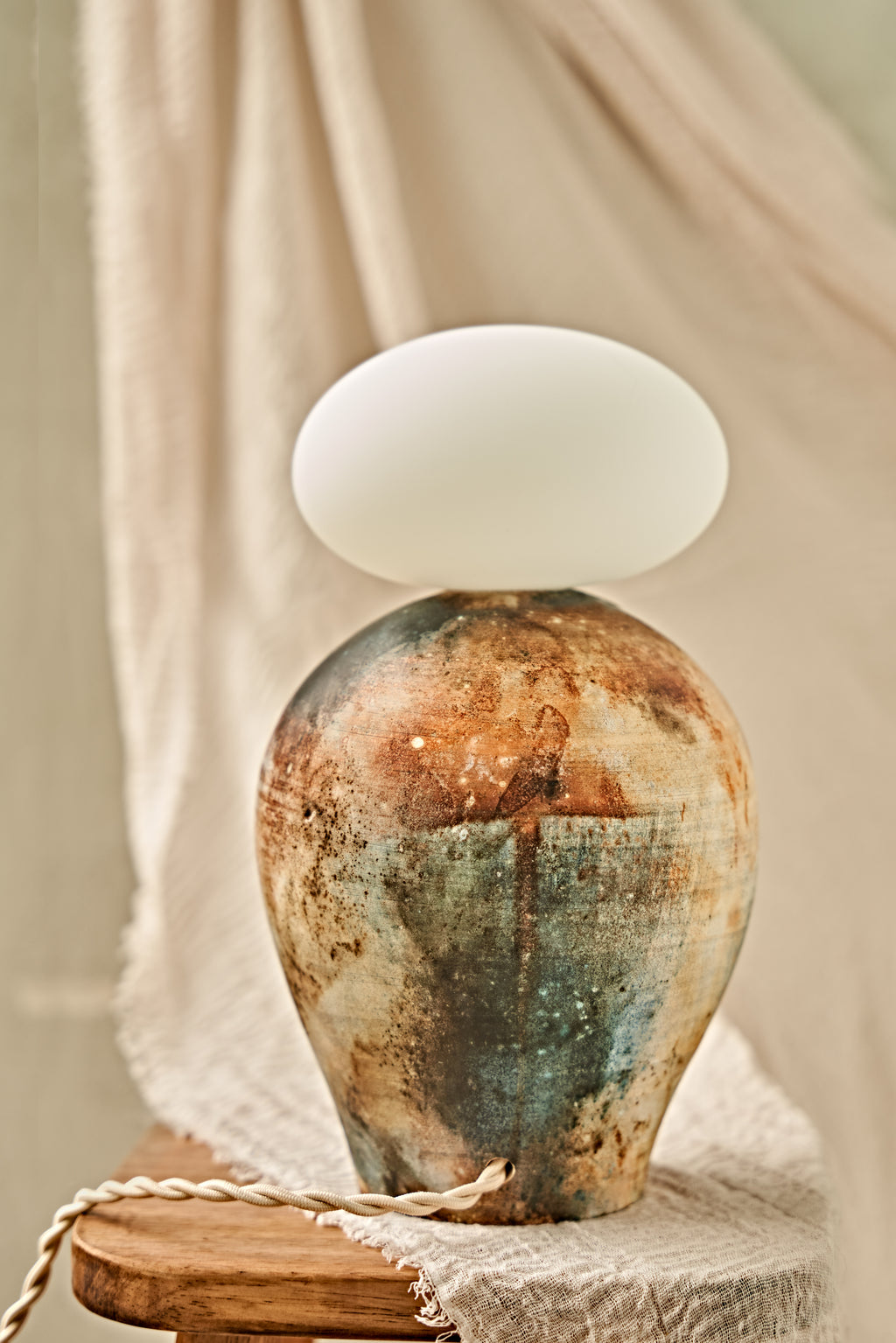 A ceramic pit-fired table lamp with a textured, earthy-toned vase body in blue, brown, and beige, topped with a glowing white oval glass orb shade, set on a wooden stool against a draped beige fabric background.