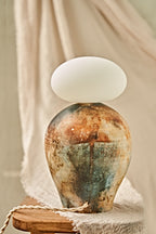 A ceramic pit-fired table lamp with a textured, earthy-toned vase body in blue, brown, and beige, topped with a glowing white oval glass orb shade, set on a wooden stool against a draped beige fabric background.
