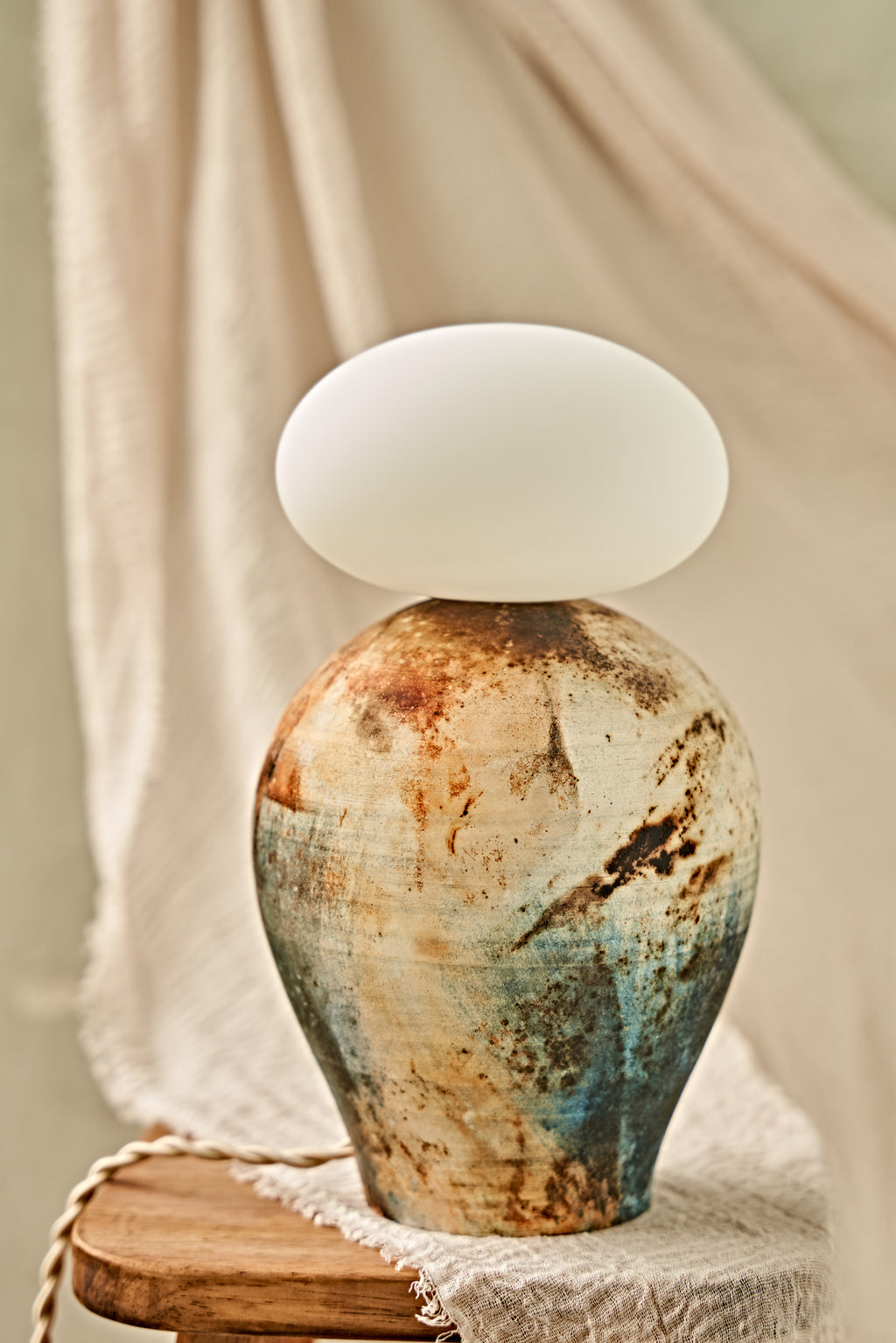 A ceramic pit-fired table lamp with a textured, earthy-toned vase body in blue, brown, and beige, topped with a glowing white oval glass orb shade, set on a wooden stool against a draped beige fabric background.