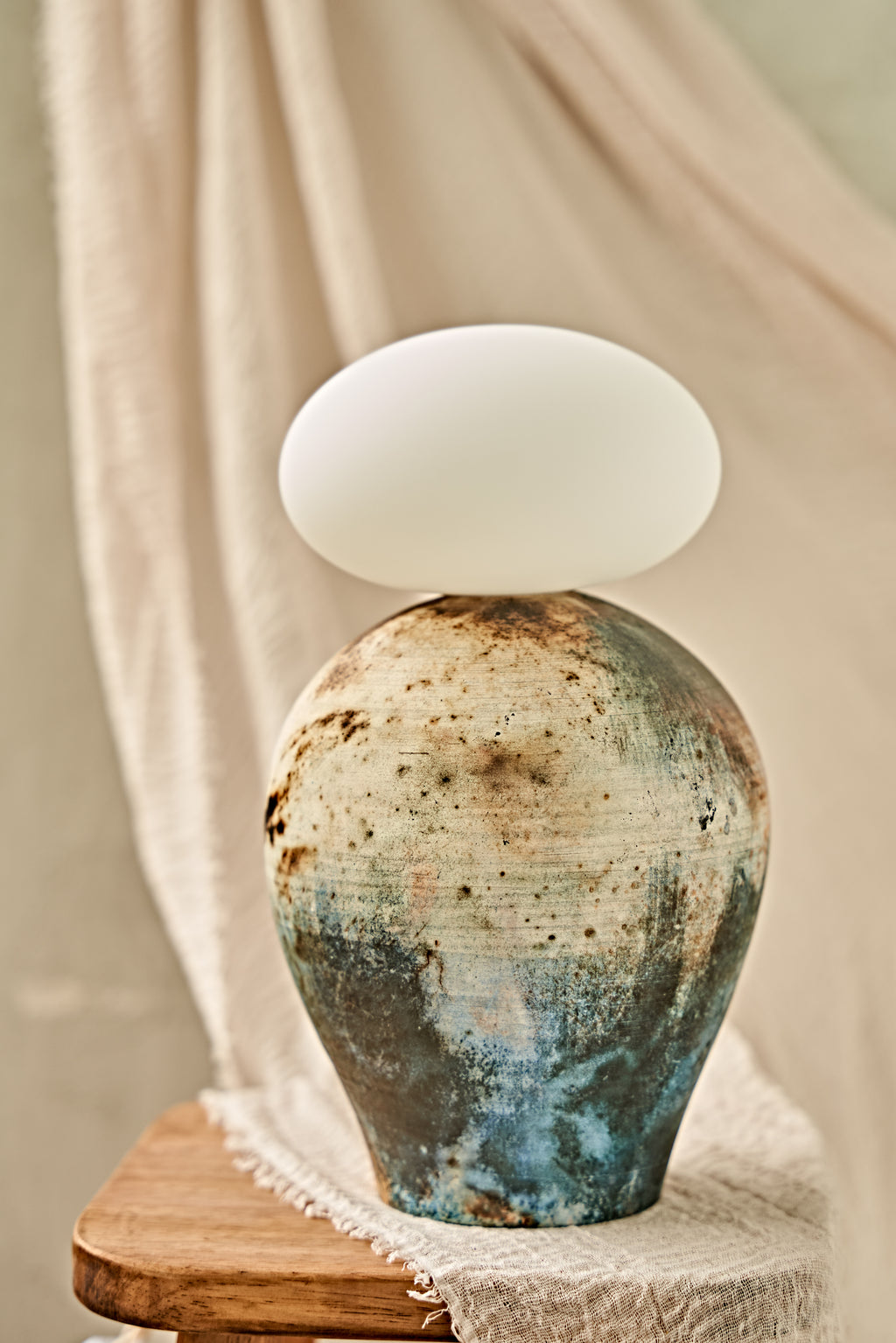A ceramic pit-fired table lamp with a textured, earthy-toned vase body in blue, brown, and beige, topped with a glowing white oval glass orb shade, set on a wooden stool against a draped beige fabric background.