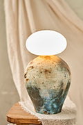 A ceramic pit-fired table lamp with a textured, earthy-toned vase body in blue, brown, and beige, topped with a glowing white oval glass orb shade, set on a wooden stool against a draped beige fabric background.