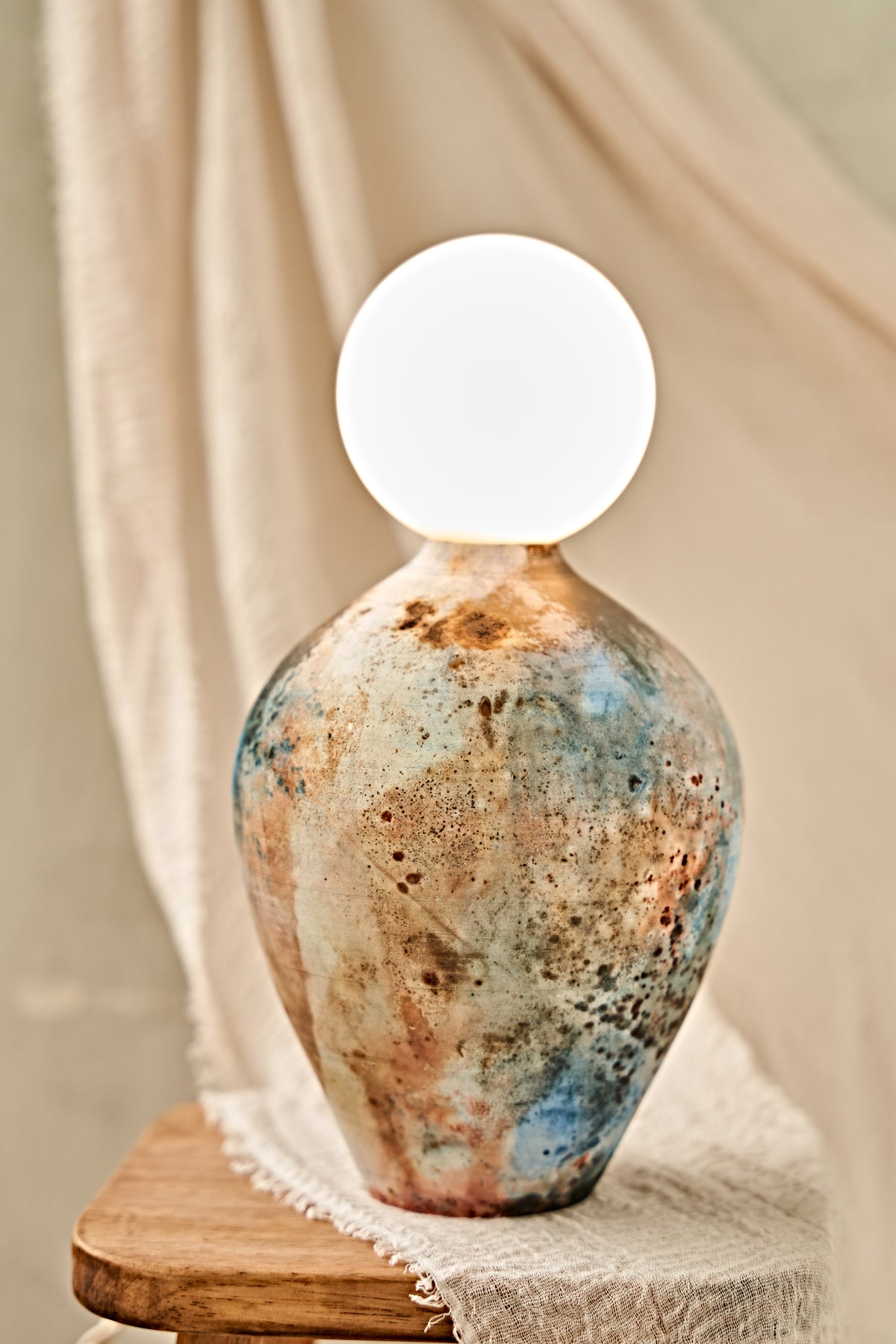 A pit-fired ceramic table lamp with a rounded, vase-like body featuring a mottled, distressed finish in earthy brown, beige, and blue tones, topped with a smooth, matte white spherical shade. The lamp is styled on a small wooden stool draped with a neutral beige fabric.