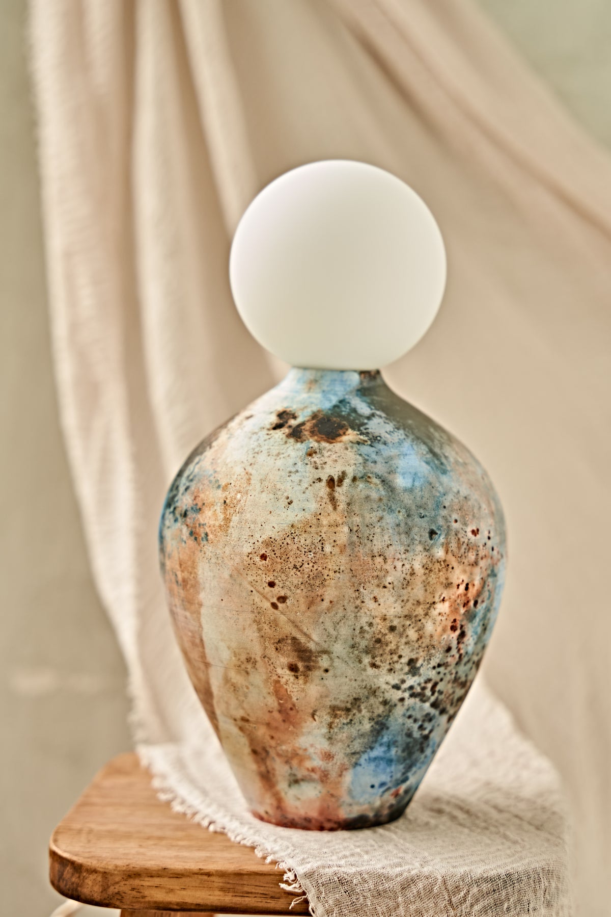 A pit-fired ceramic table lamp with a rounded, vase-like body featuring a mottled, distressed finish in earthy brown, beige, and blue tones, topped with a smooth, matte white spherical shade. The lamp is styled on a small wooden stool draped with a neutral beige fabric.
