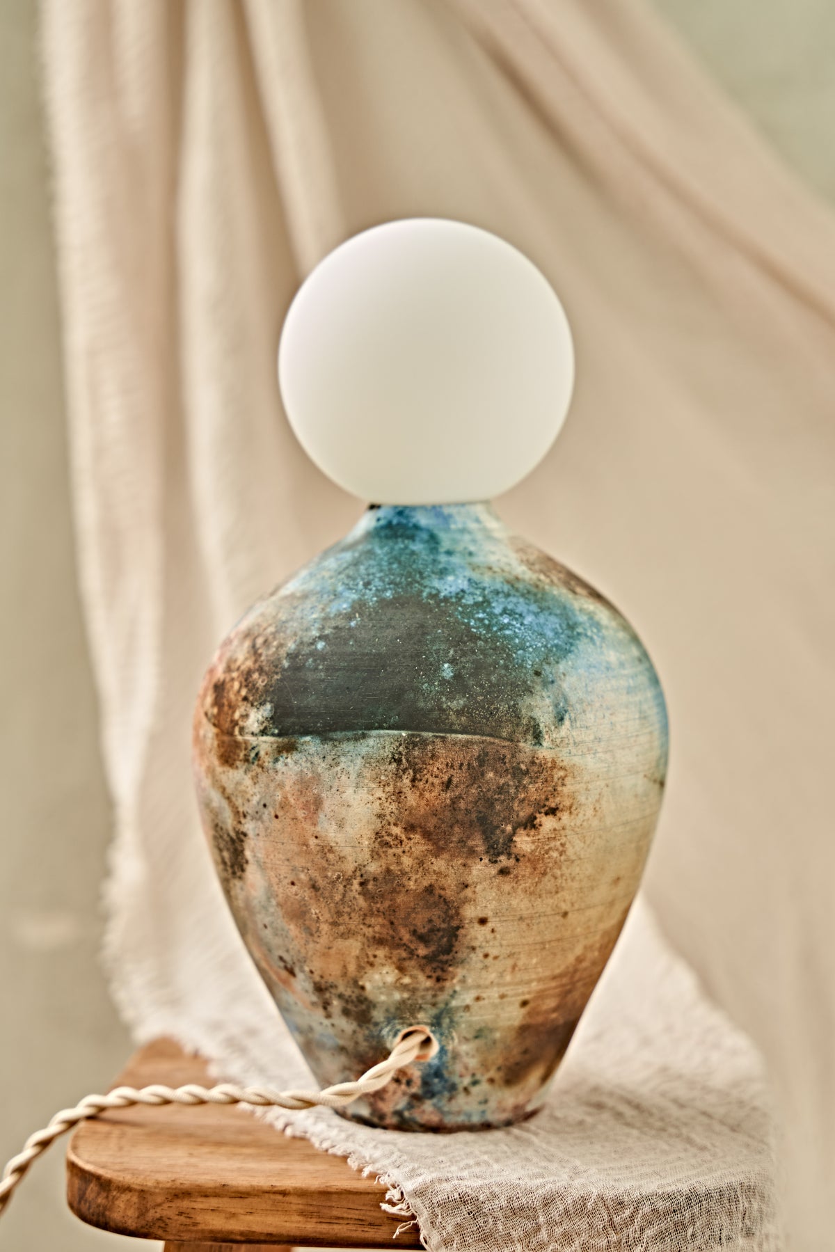 A pit-fired ceramic table lamp with a rounded, vase-like body featuring a mottled, distressed finish in earthy brown, beige, and blue tones, topped with a smooth, matte white spherical shade. The lamp is styled on a small wooden stool draped with a neutral beige fabric.