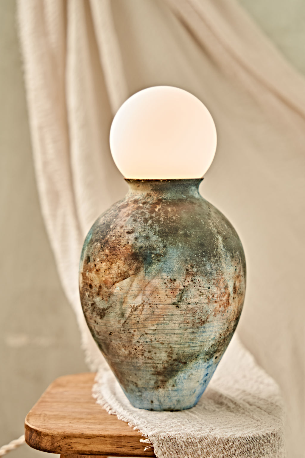 A pit-fired ceramic table lamp with a rounded, vase-like body featuring a mottled, distressed finish in earthy brown, beige, and blue tones, topped with a smooth, matte white spherical shade. The lamp is styled on a small wooden stool draped with a neutral beige fabric.