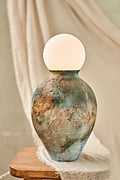 A pit-fired ceramic table lamp with a rounded, vase-like body featuring a mottled, distressed finish in earthy brown, beige, and blue tones, topped with a smooth, matte white spherical shade. The lamp is styled on a small wooden stool draped with a neutral beige fabric.