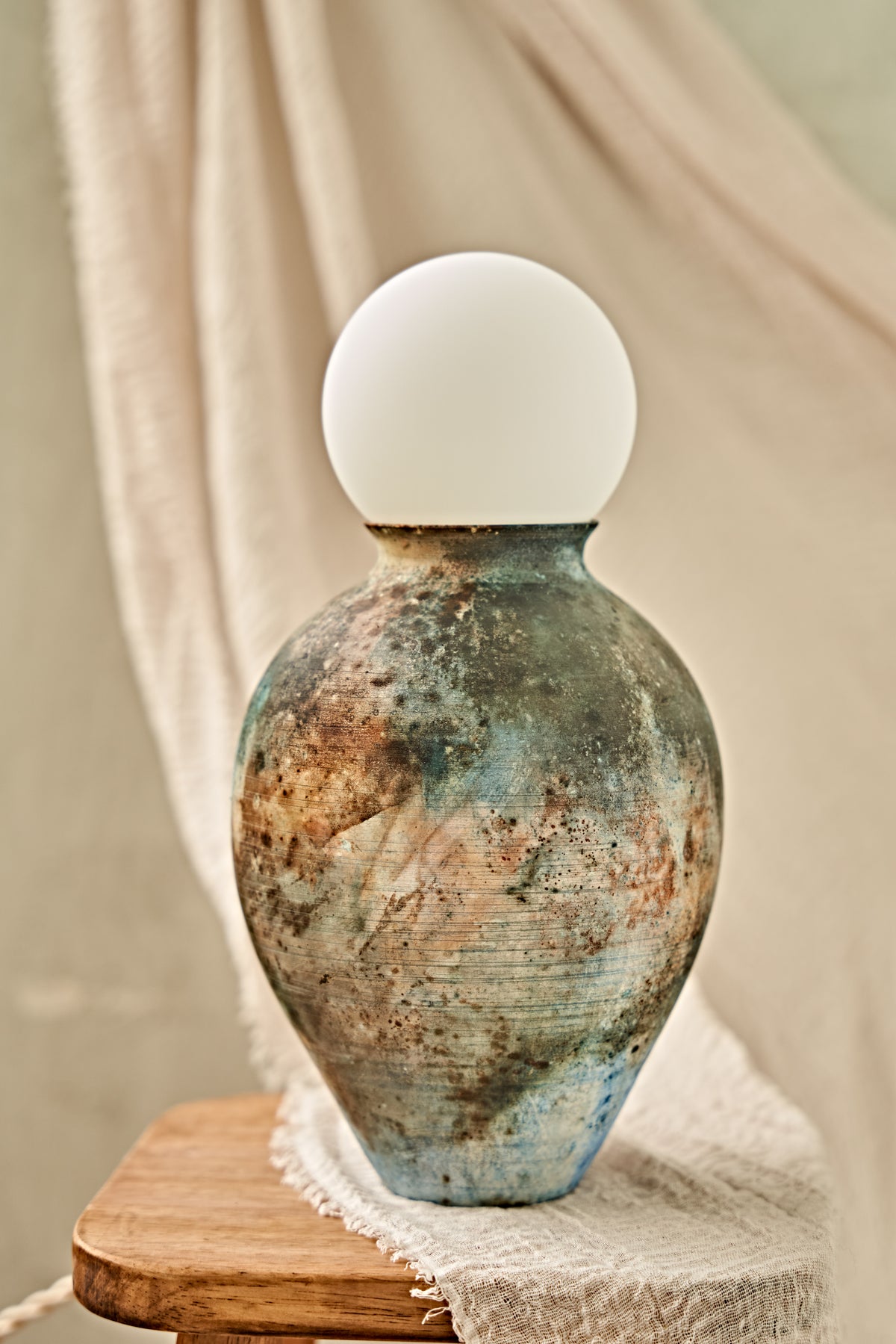 A pit-fired ceramic table lamp with a rounded, vase-like body featuring a mottled, distressed finish in earthy brown, beige, and blue tones, topped with a smooth, matte white spherical shade. The lamp is styled on a small wooden stool draped with a neutral beige fabric.