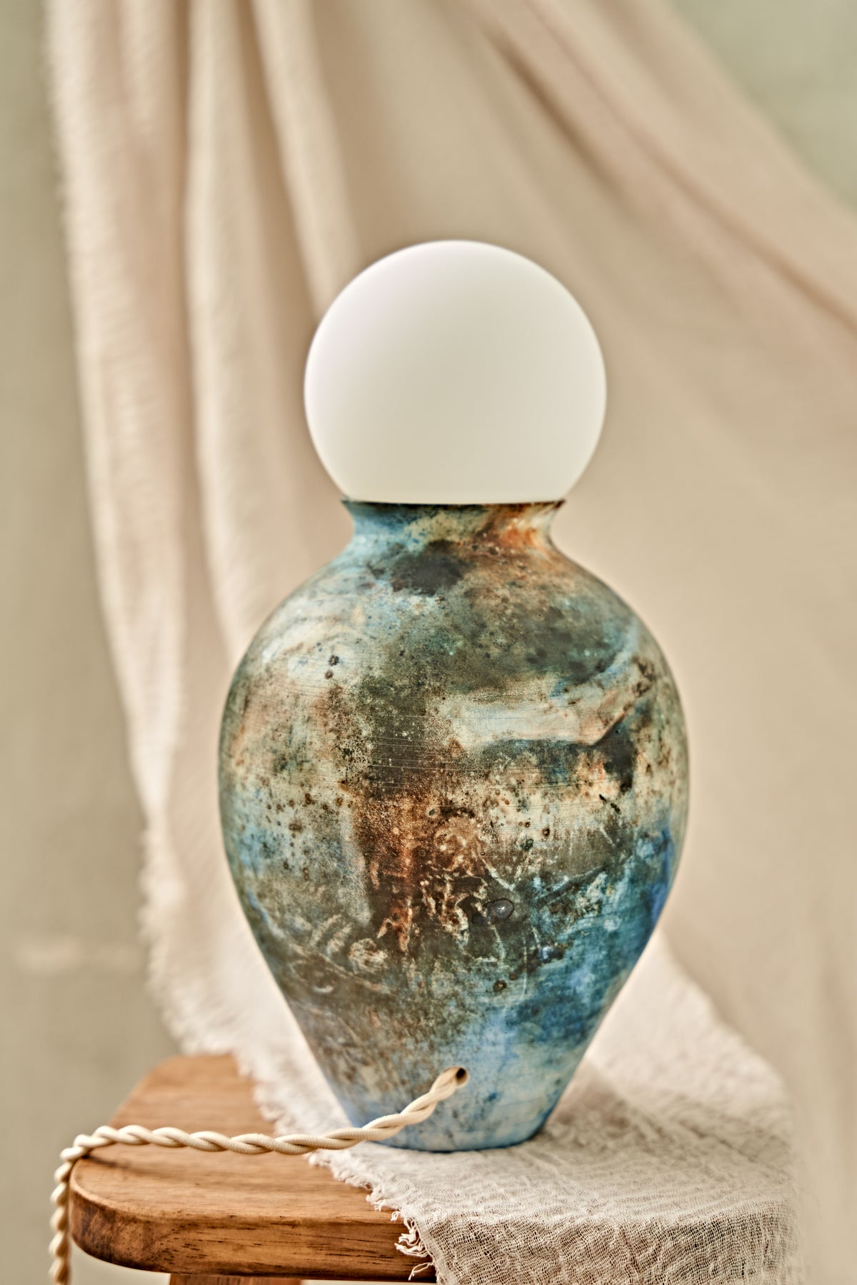 A pit-fired ceramic table lamp with a rounded, vase-like body featuring a mottled, distressed finish in earthy brown, beige, and blue tones, topped with a smooth, matte white spherical shade. The lamp is styled on a small wooden stool draped with a neutral beige fabric.