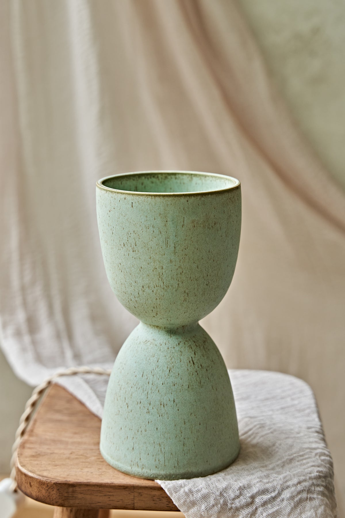 A handmade ceramic hourglass-shaped dimmable table lamp in a speckled sage green glaze with a warm yellow glow visible inside, displayed on a rustic wooden stool draped with a neutral fabric.