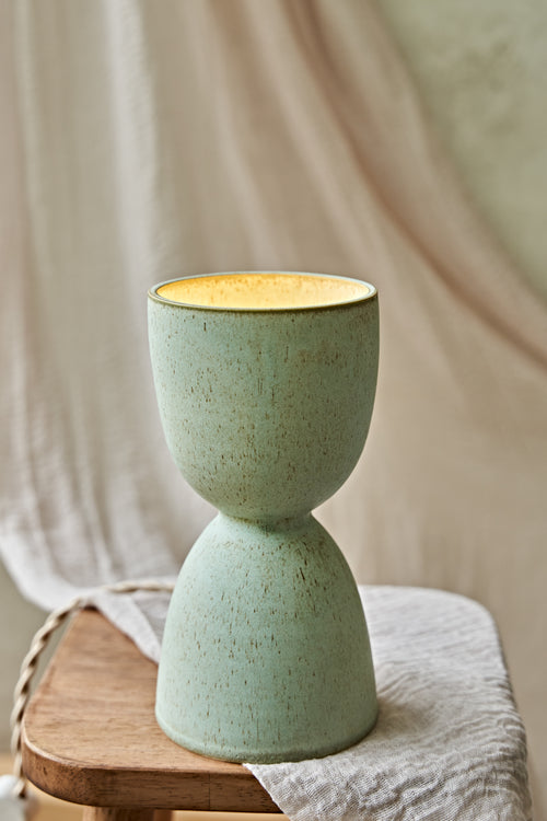 A handmade ceramic hourglass-shaped dimmable table lamp in a speckled sage green glaze with a warm yellow glow visible inside, displayed on a rustic wooden stool draped with a neutral fabric.