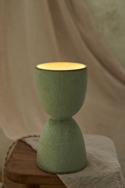 A handmade ceramic hourglass-shaped dimmable table lamp in a speckled sage green glaze with a warm yellow glow visible inside, displayed on a rustic wooden stool draped with a neutral fabric.