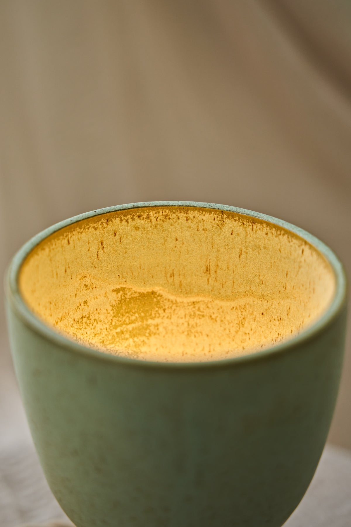 Close-up of a handmade ceramic hourglass-shaped dimmable table lamp in a speckled sage green glaze with a warm yellow glow visible inside, displayed on a rustic wooden stool draped with a neutral fabric.