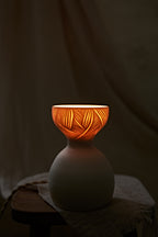 A a translucent porcelain ceramic table lamp with a matte off-white bulbous base and a flared top featuring carved leaf-like patterns that glow warm orange when lit, displayed on a rustic wooden stool with draped fabric in the background.