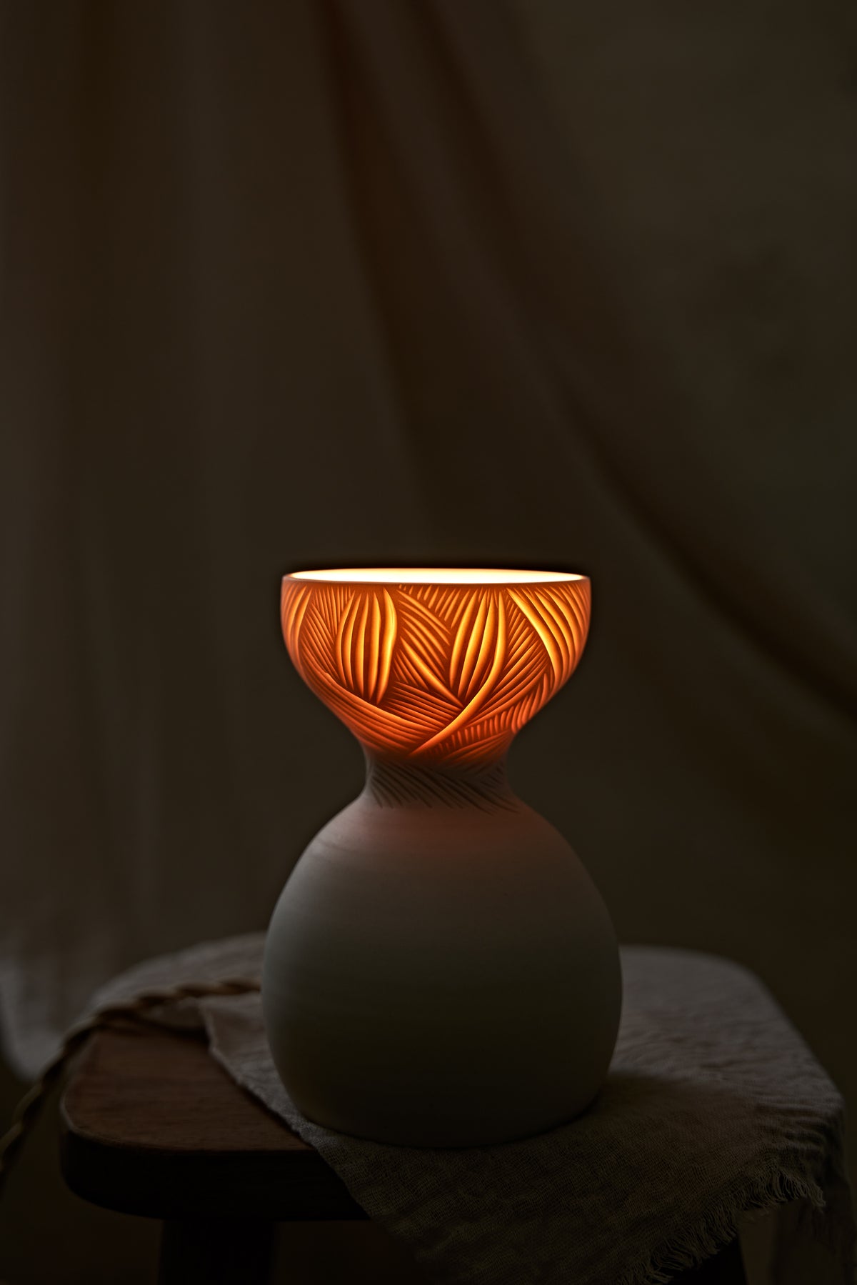 A a translucent porcelain ceramic table lamp with a matte off-white bulbous base and a flared top featuring carved leaf-like patterns that glow warm orange when lit, displayed on a rustic wooden stool with draped fabric in the background.