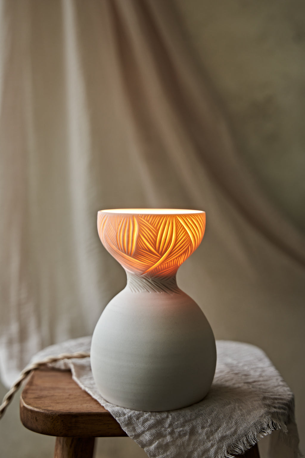 A a translucent porcelain ceramic table lamp with a matte off-white bulbous base and a flared top featuring carved leaf-like patterns that glow warm orange when lit, displayed on a rustic wooden stool with draped fabric in the background.

