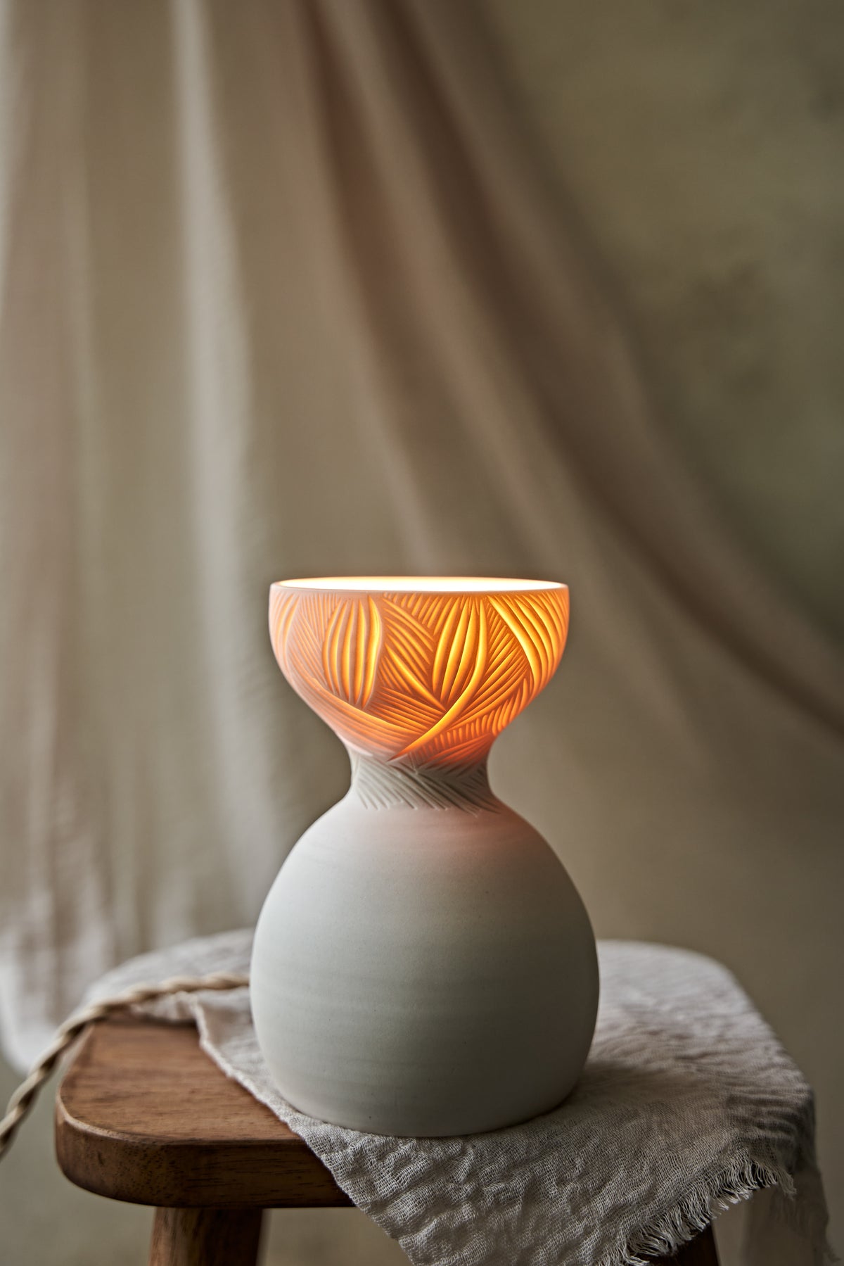 A a translucent porcelain ceramic table lamp with a matte off-white bulbous base and a flared top featuring carved leaf-like patterns that glow warm orange when lit, displayed on a rustic wooden stool with draped fabric in the background.

