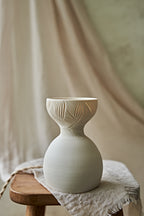 A a translucent porcelain ceramic table lamp with a matte off-white bulbous base and a flared top featuring carved leaf-like patterns that glow warm orange when lit, displayed on a rustic wooden stool with draped fabric in the background.