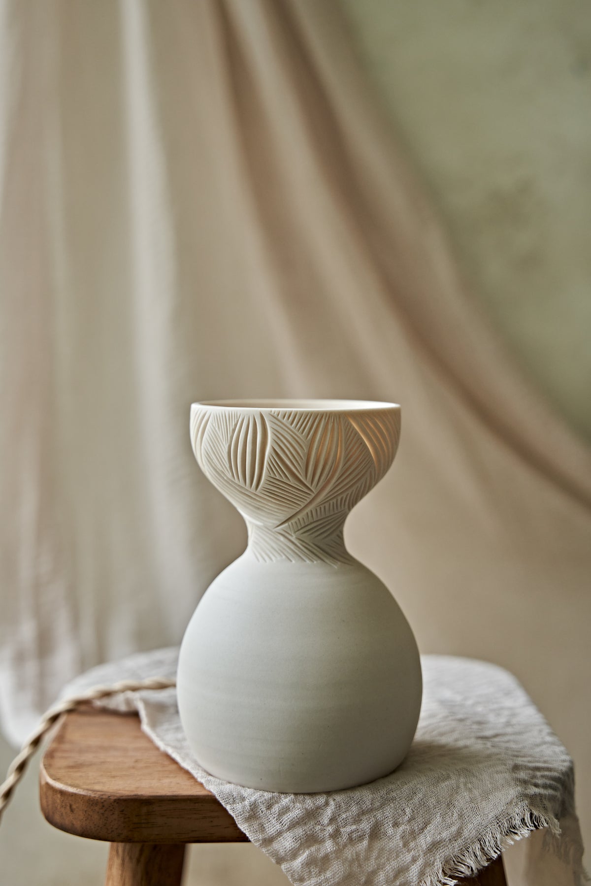 A a translucent porcelain ceramic table lamp with a matte off-white bulbous base and a flared top featuring carved leaf-like patterns that glow warm orange when lit, displayed on a rustic wooden stool with draped fabric in the background.