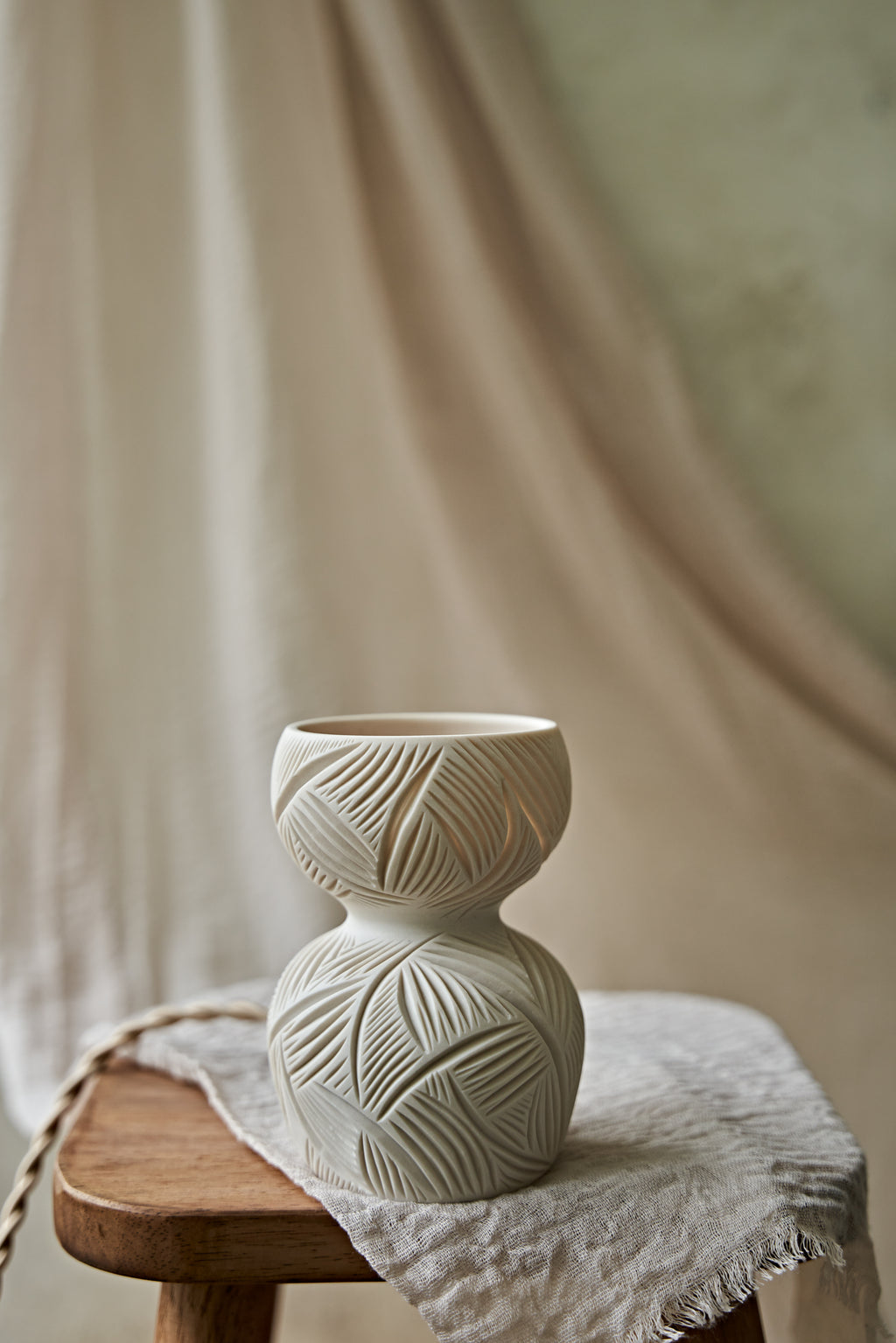 A ceramic translucent porcelain glowing lamp featuring a carved pattern on the upper half and a smooth matte white lower body, displayed on a rustic wooden stool with draped fabric in the background.