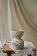 A ceramic translucent porcelain glowing lamp featuring a carved pattern on the upper half and a smooth matte white lower body, displayed on a rustic wooden stool with draped fabric in the background.