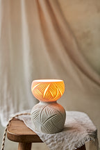 A ceramic translucent porcelain glowing lamp with a glowing warm light inside, featuring a carved pattern on the upper half and a smooth matte white lower body, displayed on a rustic wooden stool with draped fabric in the background.