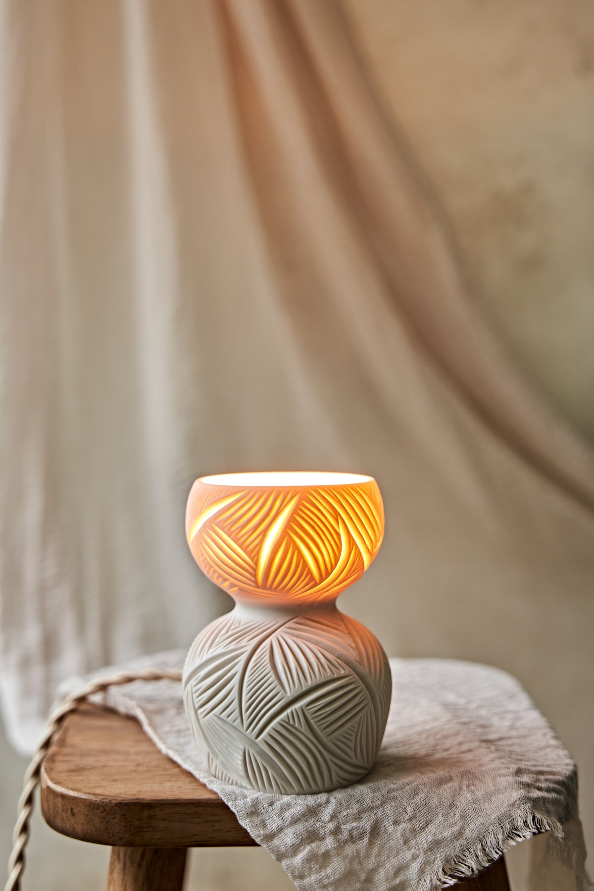 A ceramic translucent porcelain glowing lamp with a glowing warm light inside, featuring a carved pattern on the upper half and a smooth matte white lower body, displayed on a rustic wooden stool with draped fabric in the background.