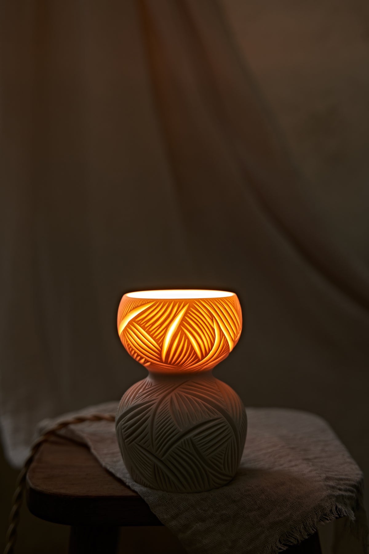 A ceramic translucent porcelain glowing lamp with a glowing warm amber light inside, featuring a carved pattern on the upper half and a smooth matte white lower body, displayed on a rustic wooden stool with draped fabric in the background.