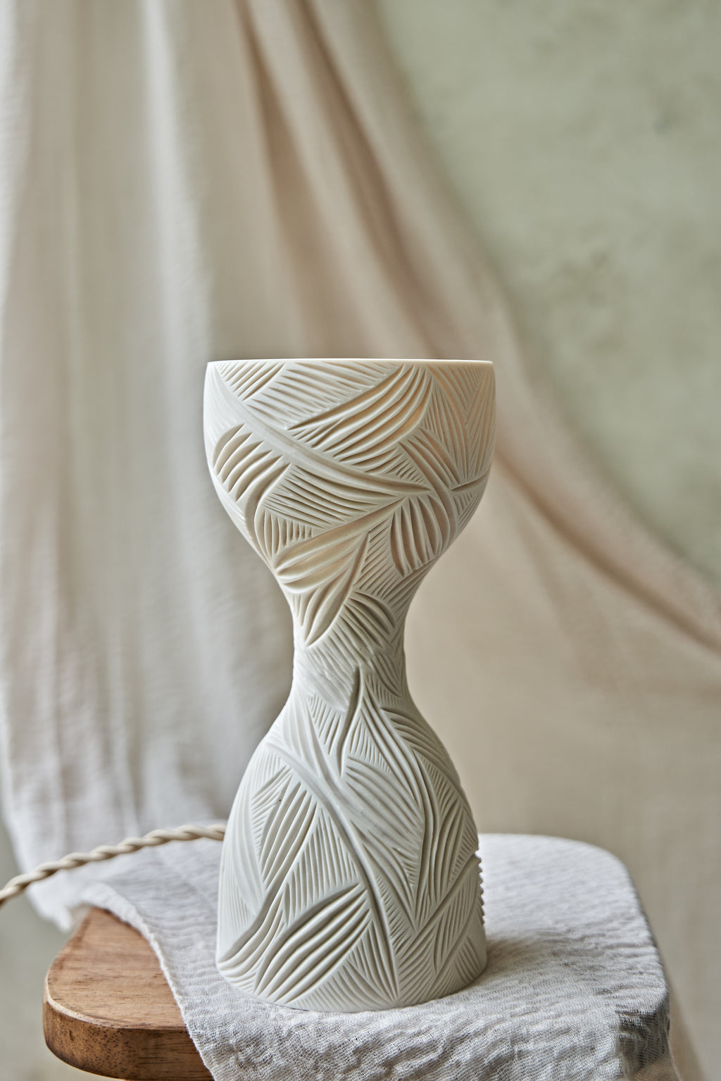 A sculptural hourglass-shaped porcelain ceramic table lamp with carved leaf patterns, glowing with warm orange light from within, set on a rustic wooden surface with draped beige fabric in the background.
