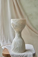 A sculptural hourglass-shaped porcelain ceramic table lamp with carved leaf patterns, glowing with warm orange light from within, set on a rustic wooden surface with draped beige fabric in the background.