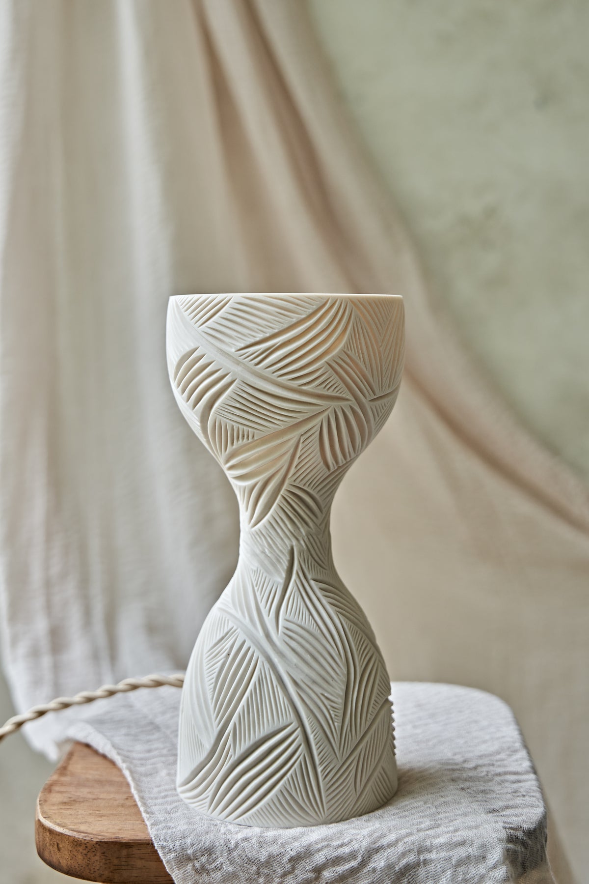 A sculptural hourglass-shaped porcelain ceramic table lamp with carved leaf patterns, glowing with warm orange light from within, set on a rustic wooden surface with draped beige fabric in the background.