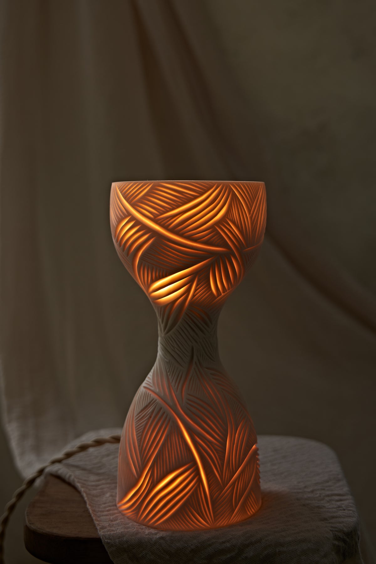 A sculptural hourglass-shaped porcelain ceramic table lamp with carved leaf patterns, glowing with warm orange light from within, set on a rustic wooden surface with draped beige fabric in the background.