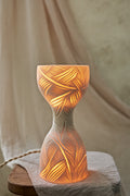 A sculptural hourglass-shaped porcelain ceramic table lamp with carved leaf patterns, glowing with warm orange light from within, set on a rustic wooden surface with draped beige fabric in the background.