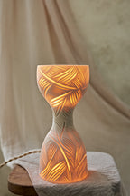 A sculptural hourglass-shaped porcelain ceramic table lamp with carved leaf patterns, glowing with warm orange light from within, set on a rustic wooden surface with draped beige fabric in the background.