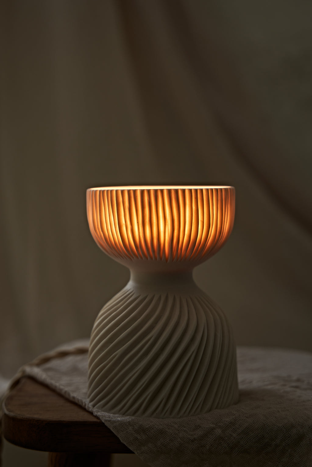 A modern translucent porcelain ceramic table lamp with a carved ribbed, flared glowing shade and a textured, ribbed base, sitting on a rustic wooden stool draped with a linen cloth.