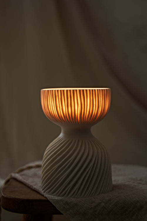 A modern translucent porcelain ceramic table lamp with a carved ribbed, flared glowing shade and a textured, ribbed base, sitting on a rustic wooden stool draped with a linen cloth.