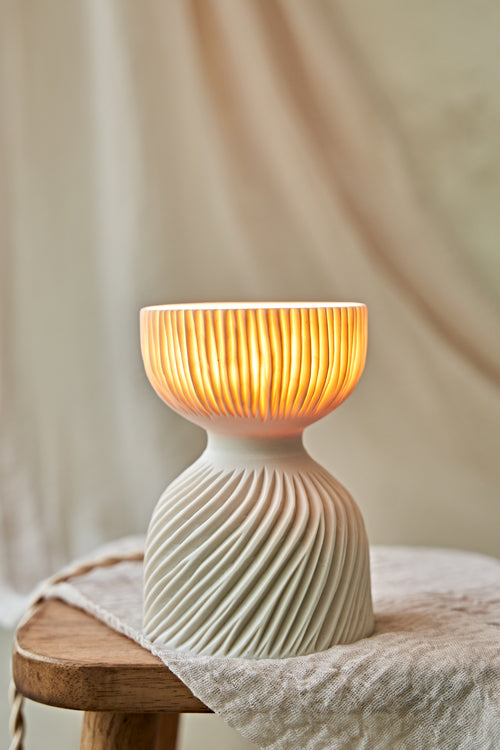 A modern translucent porcelain ceramic table lamp with a carved ribbed, flared glowing shade and a textured, ribbed base, sitting on a rustic wooden stool draped with a linen cloth.