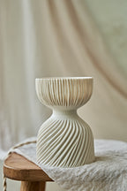 A modern translucent porcelain ceramic table lamp with a carved ribbed, flared glowing shade and a textured, ribbed base, sitting on a rustic wooden stool draped with a linen cloth.
