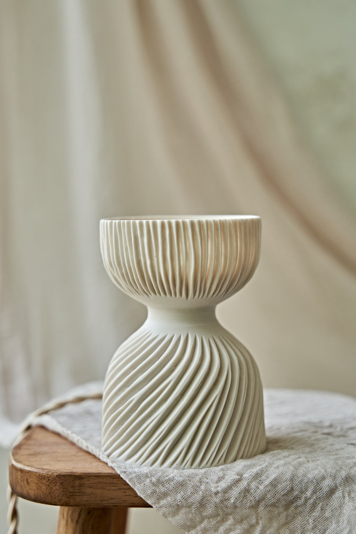 A modern translucent porcelain ceramic table lamp with a carved ribbed, flared glowing shade and a textured, ribbed base, sitting on a rustic wooden stool draped with a linen cloth.