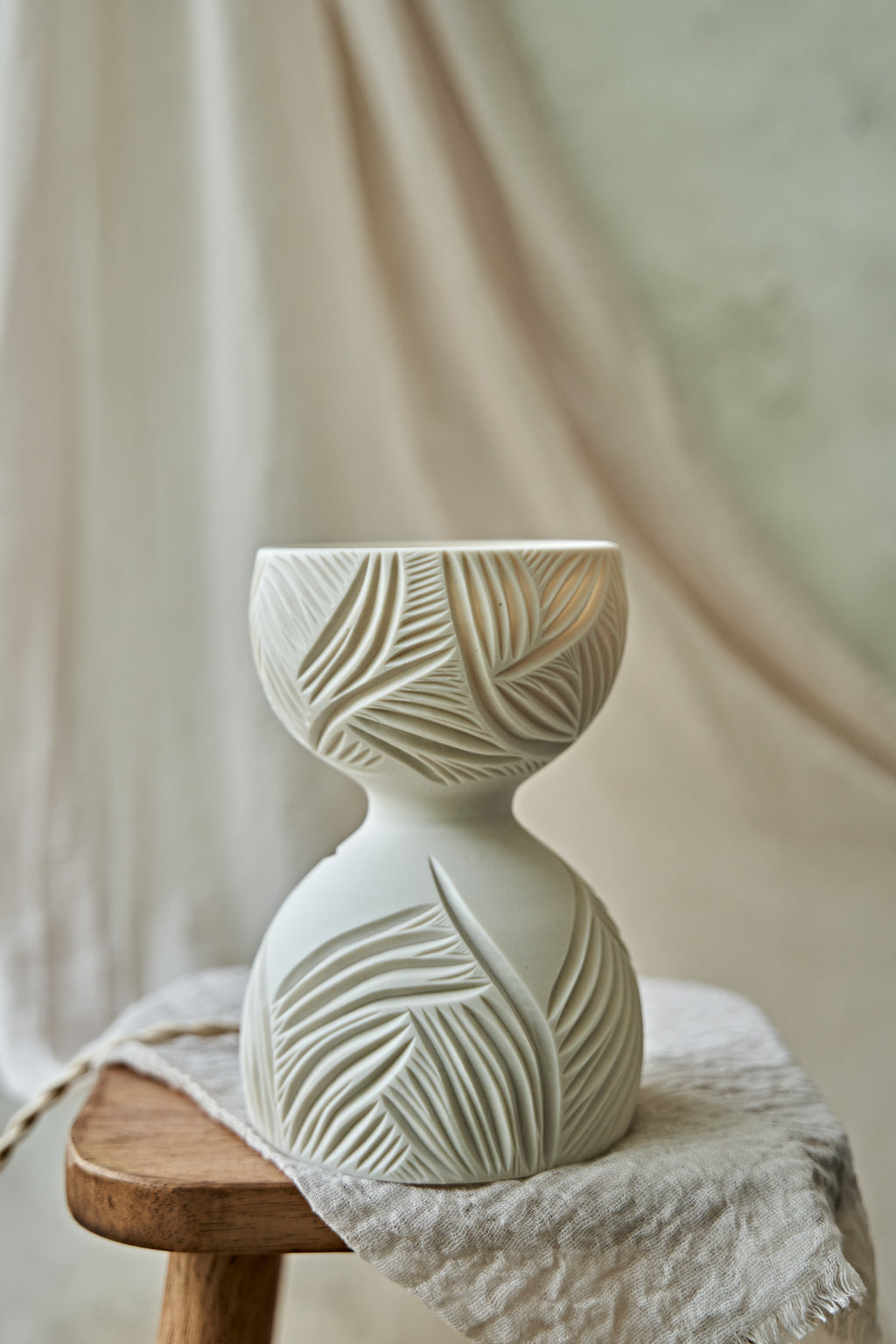 A white translucent porcelain ceramic hourglass-shaped table lamp with leaf-like carvings, glowing with warm orange light from within, displayed on a rustic wooden stool draped with a textured linen cloth against a soft beige fabric backdrop.