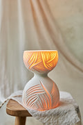 A white translucent porcelain ceramic hourglass-shaped table lamp with leaf-like carvings, glowing with warm orange light from within, displayed on a rustic wooden stool draped with a textured linen cloth against a soft beige fabric backdrop.